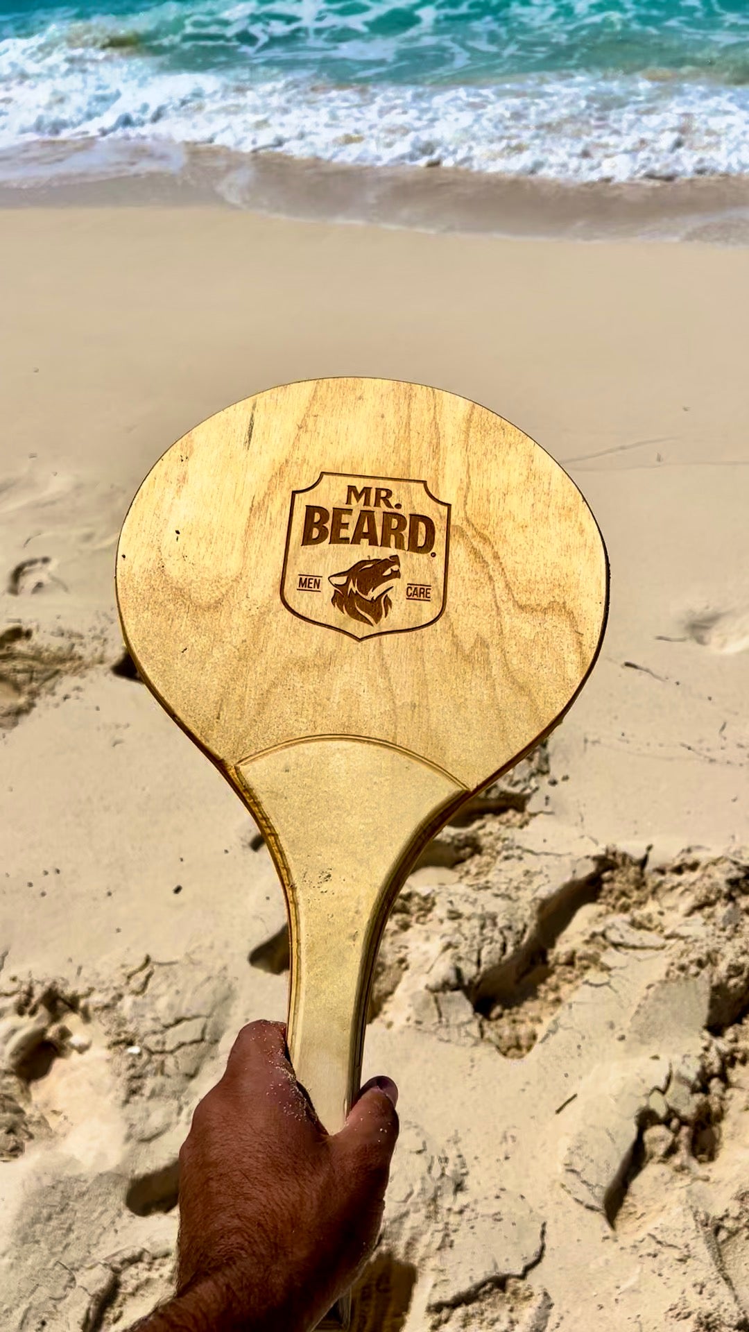 Wooden paddle with 'Mr. Beard' logo held on a sandy beach
