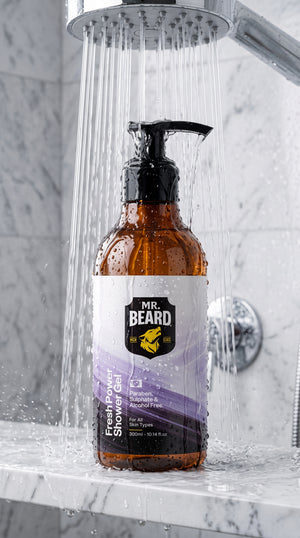 Mr. Beard Fresh Pimp Shower Gel in an amber glass pump bottle with a black dispenser, set under a running shower head on a marble countertop.