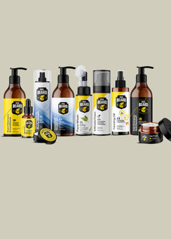 A lineup of beard care products from The Beard Brand, including pump bottles, a dropper bottle, a jar, and a small round tin, arranged against a solid light beige background.