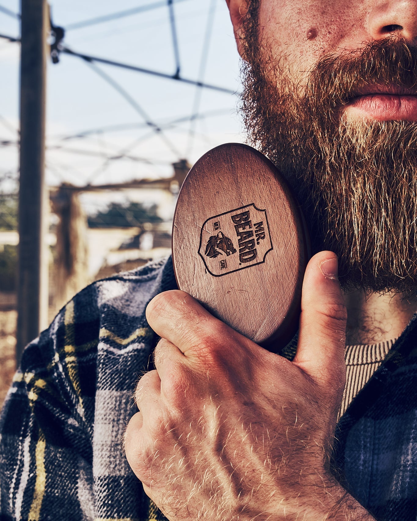 A bearded man holding a round beard brush with a wooden handle, featuring a logo.